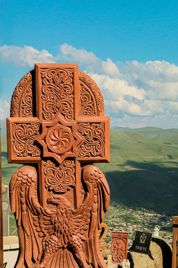hero-img-01 Detailed Armenian khachkar with intricate carvings set against a vibrant green landscape under a blue sky.